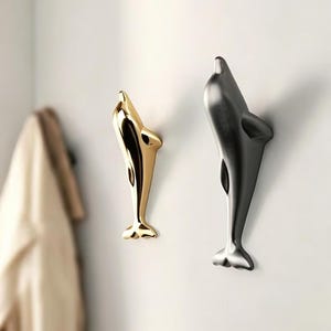 Puede incluir: Dos ganchos de pared con forma de delfín. Un gancho tiene un acabado dorado pulido y reflectante, mientras que el otro presenta un acabado gris carbón mate. Ambos ganchos están orientados verticalmente con las cabezas de los delfines hacia arriba.
