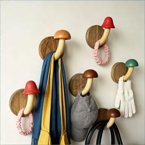 Op de afbeelding: Houten wandhaken in de vorm van paddenstoelen. De haken hebben gekleurde paddenstoelenkoppen in rood, bruin en groen. Een blauwe en gele sjaal, een grijze pet, een roze haarband en een witte handschoen hangen aan de haken.
