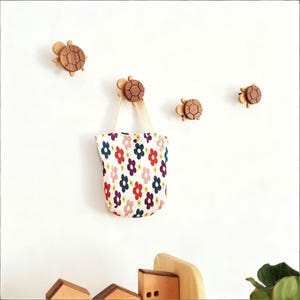 Könnte beinhalten: Wandhaken in Form von Holzschildkröten mit einer Tragetasche mit Blumenmuster, die an einem hängt. Die Haken sind aus hellbraunem Holz gefertigt und haben ein detailliertes Schildkrötenpanzer-Design. Die Tragetasche hat einen weißen Hintergrund mit bunten Blumenmustern.