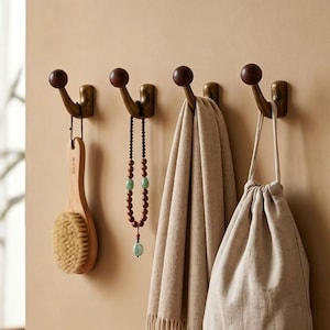May include: Four bronze-colored wall hooks with dark brown ball-shaped ends. A wooden brush, a beaded necklace, a tan scarf, and a drawstring bag hang from the hooks. The hooks are mounted on a light beige wall.