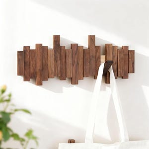 May include: A wooden wall-mounted coat rack with a unique design. The rack is made of vertical wooden slats in varying heights and widths, creating a textured surface. A white tote bag hangs from one of the hooks. The wood has a warm, natural finish.