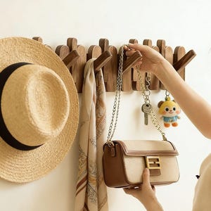 Könnte beinhalten: Eine hölzerne Wandgarderobe mit mehreren Haken. Ein Strohhut mit schwarzem Band hängt links. Ein Schal und eine Handtasche mit goldener Kette hängen ebenfalls an den Haken. Eine Person hält die Handtasche.