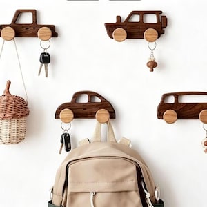May include: Four wooden car-shaped wall hooks with natural wood wheels. Each hook is a different car design. One hook holds keys, a woven basket, a backpack, and a keychain. The hooks are mounted on a white wall.