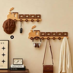 Puede incluir: Dos percheros de pared de madera con forma de tren, con una locomotora de madera clara y vagones de madera oscura. Cada tren tiene varios ganchos circulares para colgar objetos como una gorra marrón, un llavero, auriculares, un bolso y una tela blanca.