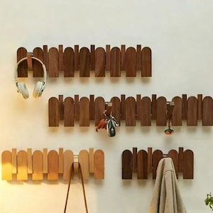 May include: A selection of wooden fence-style wall hooks in shades of brown, mounted on a white wall. Items including headphones, keys, a bag, and a towel are hung from the hooks. The design is both decorative and practical.