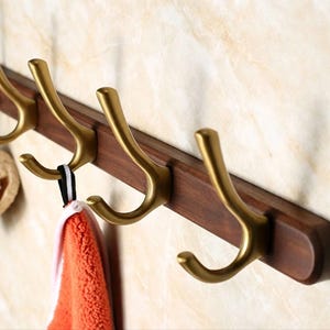 May include: A wall-mounted coat rack featuring a dark brown wooden horizontal base with four attached matte gold metal hooks. Each hook has a curved, dual-pronged design. An orange textured towel hangs from one of the metal hooks.