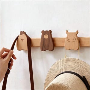 May include: A wall-mounted wooden rack featuring three distinct animal-shaped hooks. The left hook is light wood, the center is dark walnut wood shaped like a bear, and the right is light wood shaped like a cow, holding a straw hat.