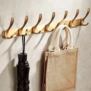 May include: A wall-mounted coat rack featuring a long, horizontal brushed gold metal bar with seven upward-curving hooks. A black umbrella hangs from the first hook, and a beige woven fabric tote bag hangs from the sixth hook on the rack.