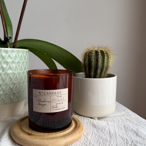 May include: A cylindrical amber glass candle jar with a cream-coloured label featuring the text V CANDLES, Strawberry Mango, 19/12/25, and Victor. The candle sits on a round, light-coloured wooden coaster, positioned between a green plant and a cactus.