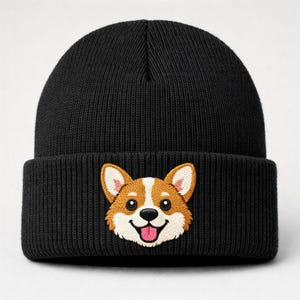 May include: Black knit beanie featuring an embroidered corgi face. The corgi has brown, white, and pink details, with a smiling expression. The beanie has a folded cuff and is made of a ribbed knit material.