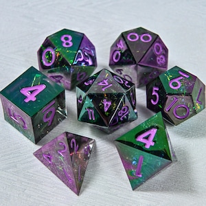 May include: A collection of seven iridescent polyhedral dice with purple numerals. The dice feature a green and purple colour scheme, with a shimmering, speckled appearance. The dice are designed for tabletop gaming.
