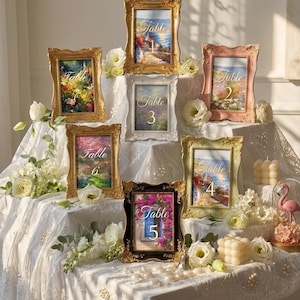 May include: Ornate picture frames with landscape paintings and table numbers. The frames are gold, pink, and black, and the paintings depict scenes of flowers, gardens, and coastal views. White flowers and candles decorate the table.