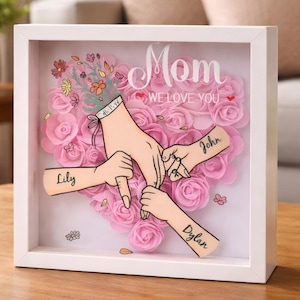 Op de afbeelding: Een wit omkaderde schaduwdoos met een roze rozenhartontwerp. De woorden "Mom WE LOVE YOU" staan bovenaan. De doos bevat illustraties van handen die elkaar vasthouden, met de namen "Lily", "John" en "Dylan".
