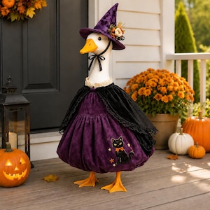 May include: A white goose wearing a purple velvet witch hat with floral accents, a black lace-trimmed cape, and a purple velvet dress featuring an embroidered black cat and orange stars. The goose has a bright orange beak and orange webbed feet.