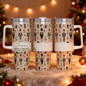 May include: Three stainless steel travel mugs with white handles and lids. The mugs feature a beige background with a repeating pattern of nutcracker soldiers and floral accents. The text "Jason" and "Merry Christmas and Happy New Year" is printed on the front.