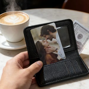 May include: Black leather wallet with a textured finish, holding a photo card with a couple kissing. A cup of coffee with latte art and a dollar bill are visible in the background.