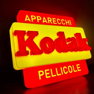 Può includere: Un'insegna luminosa rettangolare con il logo Kodak rosso in rilievo su sfondo giallo. Sopra il logo è stampata la parola APPARECCHI in giallo, mentre sotto il logo è presente la parola PELLICOLE stampata in giallo su sfondo rosso.