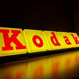 Può includere: Un'insegna rettangolare illuminata che presenta la parola Kodak in lettere rosse in grassetto, maiuscole e minuscole. Ogni lettera è alloggiata all'interno di un singolo blocco quadrato giallo, creando un effetto tridimensionale luminoso e brillante.