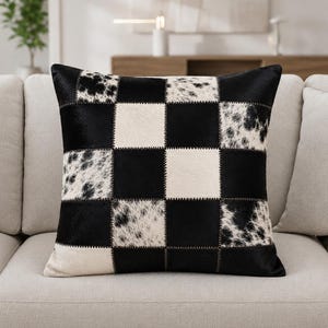 May include: A square throw pillow featuring a checkerboard pattern made of alternating patches of solid black leather and white cowhide with black spots. The individual squares are joined together with visible dark stitching throughout.