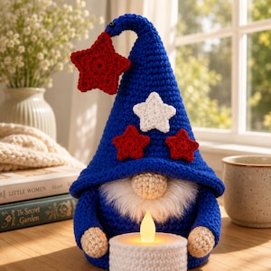 May include: A crochet gnome in patriotic colours, featuring a blue hat decorated with red and white stars. The gnome has a white beard, beige nose and hands, and holds a white candle holder with a glowing LED candle. A decorative item.