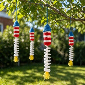May include: Crocheted rocket ornaments in red, white, and blue hang from a tree branch. The rockets have a blue cone-shaped top, red and white striped body, and a white, zig-zagging tail with yellow fringe. The ornaments are suspended by blue strings.