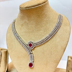 May include: A silver-toned necklace featuring a double-row leaf pattern set with numerous small clear stones. The design includes a circular accent piece with a central round red stone and a dangling teardrop-shaped red stone at the bottom.