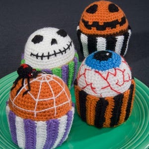 Può includere: Quattro decorazioni all'uncinetto a forma di cupcake con temi di Halloween. Una è un teschio bianco, una è una zucca arancione, una è un bulbo oculare bianco con vene rosse e una ha una parte superiore arancione con una ragnatela bianca e un ragno nero.