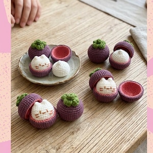 Könnte beinhalten: Eine Sammlung von gehäkelten Mangostanfrüchten in Lila und Grün, einige mit kleinen weißen katzenartigen Figuren im Inneren. Die Früchte haben texturierte, kugelförmige Formen mit abnehmbaren Deckeln, die eine weiße Katze oder ein rosa Inneres zeigen.