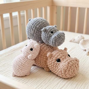 Könnte beinhalten: Drei Plüsch-Nilpferde aus grobem, weichem Garn. Ein Nilpferd ist hellgrau, eines ist hellrosa und das größte ist beige. Jedes Spielzeug hat einen abgerundeten Körper, kleine Ohren und schwarze Kunststoffaugen. Sie sind aufeinandergestapelt.