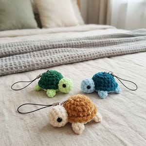Könnte beinhalten: Drei kleine gehäkelte Schildkröten-Schlüsselanhänger mit schwarzen Perlenaugen und schwarzen Kordelschlaufen. Eine Schildkröte hat einen dunkelgrünen Panzer und hellgrünen Körper, eine einen blaugrünen Panzer und hellblauen Körper, eine einen braunen Panzer.