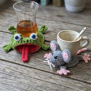 Könnte beinhalten: Ein grüner gehäkelter Frosch-Untersetzer mit roter Zunge und blauen Augen unter einem Teeglas sowie ein grauer gehäkelter Maus-Untersetzer mit rosa Ohren und Pfoten unter einer kleinen beigen Keramiktasse mit einem weißen Löffel.