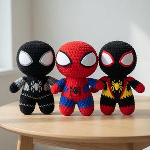 May include: Three handmade crochet dolls featuring large round heads and white oval eyes. The left doll is black with silver accents, the centre doll is red and blue, and the right doll is black, red, and yellow with a prominent spider emblem.