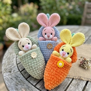 Könnte beinhalten: Drei gehäkelte Plüschtiere in Form kleiner Hasen, die in strukturierten Beuteln stecken. Ein cremefarbener Hase in einem salbeigrünen Beutel, ein rosa Hase in einem blauen und ein gelber in einem orangefarbenen. Jeder Beutel hat eine kleine Blüte.