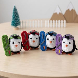 Könnte beinhalten: Vier kleine gehäkelte Pinguinfiguren, die jeweils eine passende Mütze tragen und ein entsprechendes Snowboard halten. Die Pinguine haben weiße Gesichter, schwarze Körper und orangefarbene Schnäbel. Die Snowboards sind grün, rot, blau und lila.