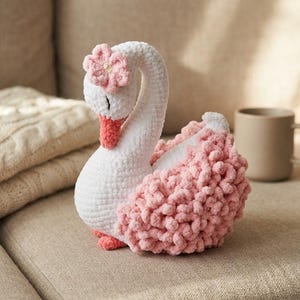 Könnte beinhalten: Ein Plüschschwan aus weißem und zartrosa Garn. Der Schwan hat einen langen, geschwungenen Hals, einen korallenfarbenen Schnabel und eine kleine rosa Blumenverzierung auf dem Kopf. Die Flügel bestehen aus strukturiertem, geschlungenem rosafarbenem Garn.
