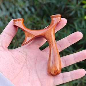 May include: A handcrafted wooden slingshot with a polished finish. The slingshot features a Y-shaped frame with rounded edges and a smooth, ergonomic handle. The wood grain is visible, showcasing the natural color and texture of the material.