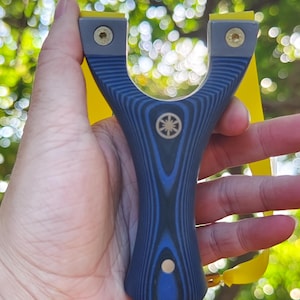 Op de afbeelding: Een blauwe en grijze katapult met een houtnerfpatroon. De katapult heeft gele banden en messing hardware. Een klein cirkelvormig embleem is gecentreerd op het handvat.