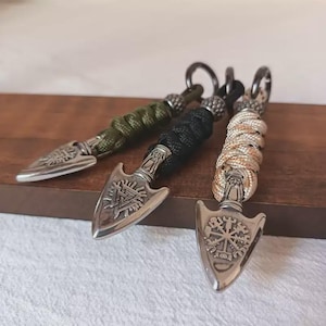 Puede incluir: Tres llaveros con puntas de flecha de metal plateado unidas a mangos de paracord trenzado. Las puntas de flecha muestran intrincados símbolos en relieve, incluyendo un Valknut y un Vegvisir. Cada mango está asegurado con una cuenta y una anilla.