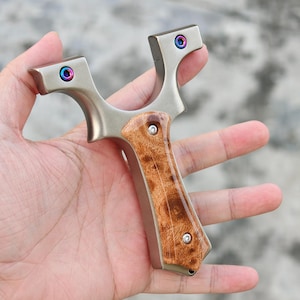 Könnte beinhalten: Eine silberne Steinschleuder mit Holzgriff. Die Steinschleuder hat einen Metallrahmen mit farbigen Akzenten auf der Oberseite. Der Griff besteht aus Holz mit einem natürlichen Maserungsmuster. Die Steinschleuder wird in einer Hand gehalten.