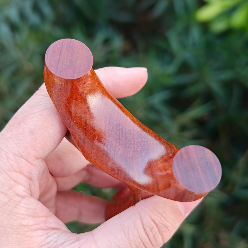 May include: A handcrafted wooden slingshot with a curved, polished body and two circular end pieces. The slingshot is made of reddish-brown wood, showcasing the natural grain and texture. The design is held in a hand against a blurred green background.