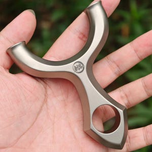 Könnte beinhalten: Eine metallisch graue Steinschleuder mit einem Y-förmigen Rahmen und einem kreisförmigen Loch am Griff. Die Steinschleuder hat eine glatte, matte Oberfläche und ein kleines kreisförmiges Emblem mit japanischen Schriftzeichen.