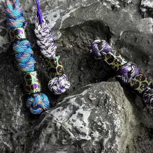 May include: Close-up of three handmade paracord accessories. The accessories feature colourful, knotted cords in blue, purple, and white. Each accessory has decorative metal beads with a skull design. The background is a textured, grey stone surface.