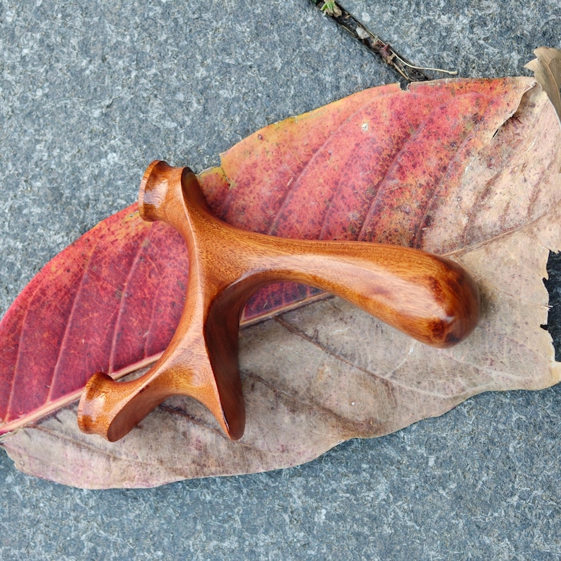 May include: A handcrafted wooden slingshot with a smooth, curved handle and a forked frame. The slingshot is made of polished wood, showcasing its natural grain and color. It rests on a red and brown leaf against a gray stone surface.