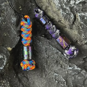 Op de afbeelding: Twee paracord-lanyards met gevlochten knopen en cilindrische kralen. De ene lanyard heeft een felgekleurd regenboogkoord met een donkere kraal met een groene schedelafbeelding. De andere heeft een paars en zwart koord met een donkere kraal met patroon.
