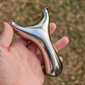 May include: A polished, chrome-like slingshot with a Y-shaped frame and a rounded handle. The slingshot is held in a hand, showcasing its smooth, reflective surface. The design suggests a classic, recreational item.