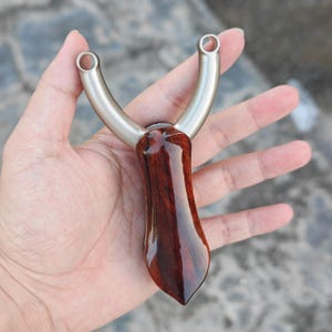 Könnte beinhalten: Eine Steinschleuder mit einem polierten, dunkelrotbraunen Holzgriff und einem silberfarbenen, gebogenen Metallrahmen. Der Griff hat eine glänzende Oberfläche, und der Metallrahmen hat kreisförmige Öffnungen an der Oberseite.
