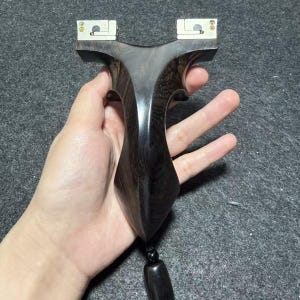 Könnte beinhalten: Eine dunkle Holzschleuder mit glatter, polierter Oberfläche. Die Schleuder hat einen Y-förmigen Rahmen mit Metallbändern oben zur Befestigung der Bänder. Eine schwarze Perle und ein rechteckiges Holzstück sind unten befestigt.