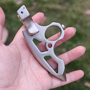 May include: A silver-colored, multi-tool knife with a finger loop and a pocket clip. The knife has a pointed blade and a unique, open design. The tool is held in a hand against a blurred green background.