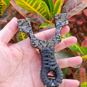 Könnte beinhalten: Eine schwarze und silberne Steinschleuder mit floralem Design. Die Steinschleuder hat einen geflochtenen schwarzen Griff. Der Rahmen ist dunkel mit einem detaillierten Blumenmuster. Die Steinschleuder wird in einer Hand vor einem Hintergrund aus bunten Blättern gehalten.