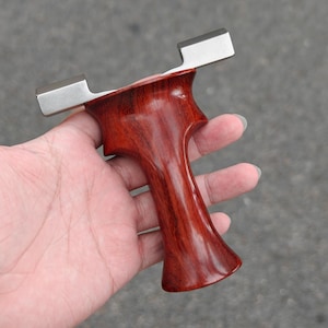Könnte beinhalten: Eine Schleuder mit einem polierten, rotbraunen Holzgriff und einem silberfarbenen Metallrahmen. Der Griff hat eine konturierte Form für einen bequemen Griff. Die Schleuder ist für den Freizeitgebrauch konzipiert.